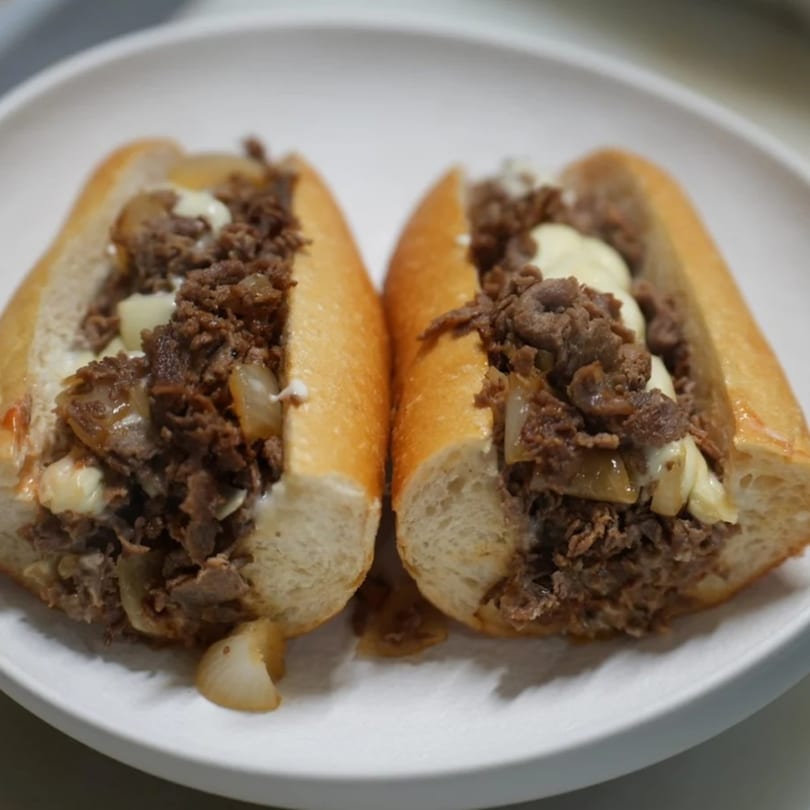 Two delicious cheesesteak sandwiches with steak, onions, and melted cheese on a white plate.
